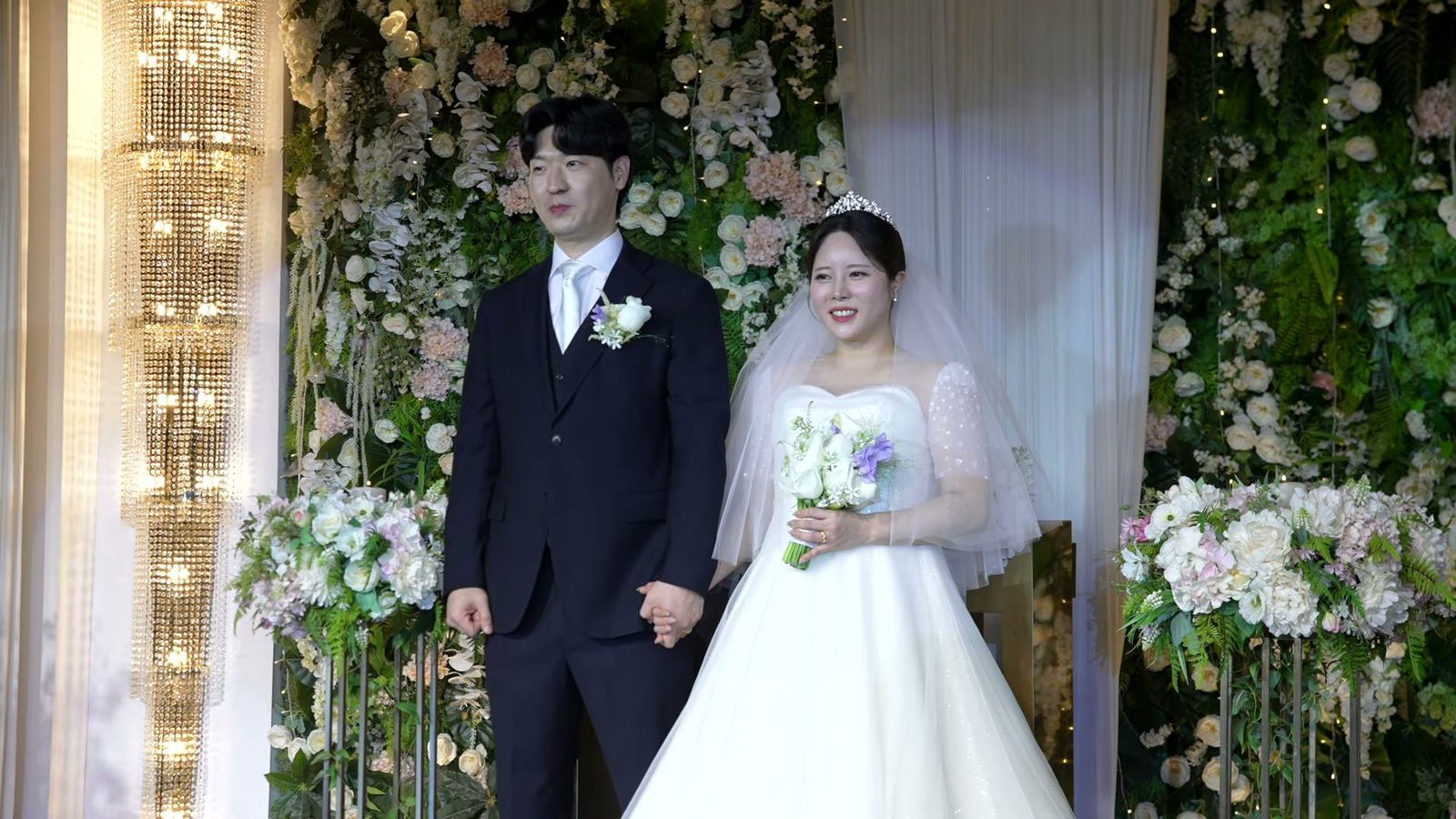 초록 잎과 흰 꽃으로 덮인 꽃벽 무대 앞에서 손을 잡고 선 신랑 신부, 왼쪽에 크리스털 샹들리에