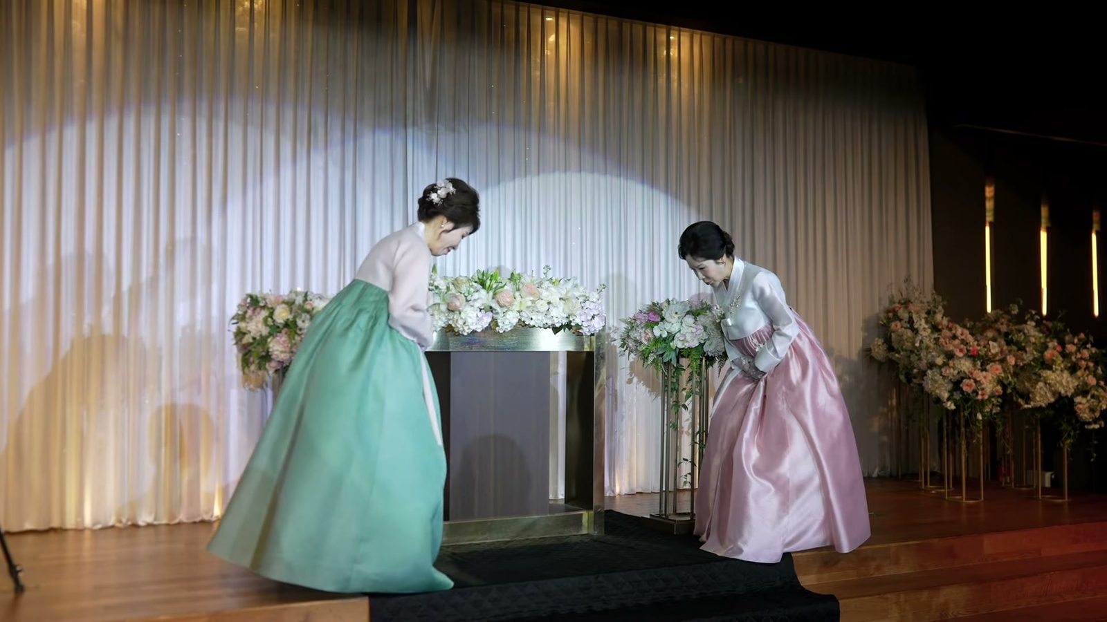 흰 주름 커튼과 나무 바닥 무대에서 금색 테두리 탁자를 사이에 두고 고개 숙여 인사하는 한복 차림의 두 어머님
