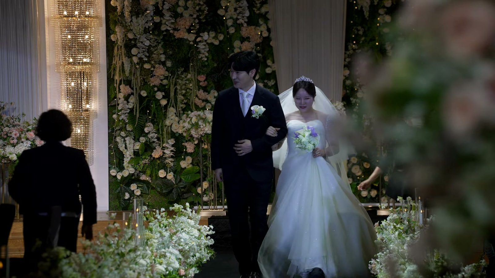 흰 꽃과 유리 촛대가 놓인 버진로드를 팔짱 끼고 걸어 나오는 신랑 신부, 뒤로 꽃벽