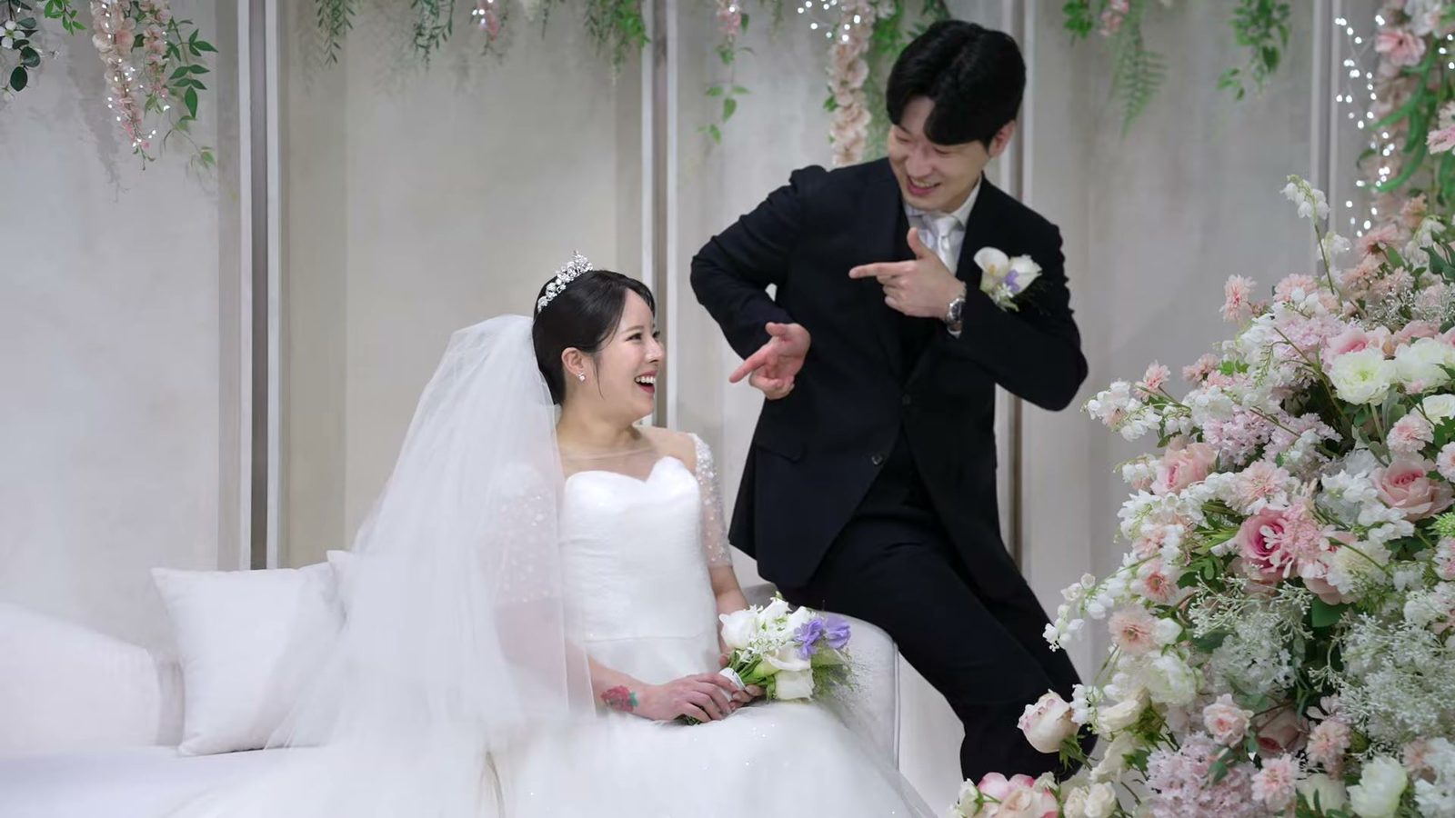흰 소파에 앉아 웃는 신부와 옆에서 장난스럽게 손가락으로 가리키는 신랑, 위로 늘어진 분홍 꽃