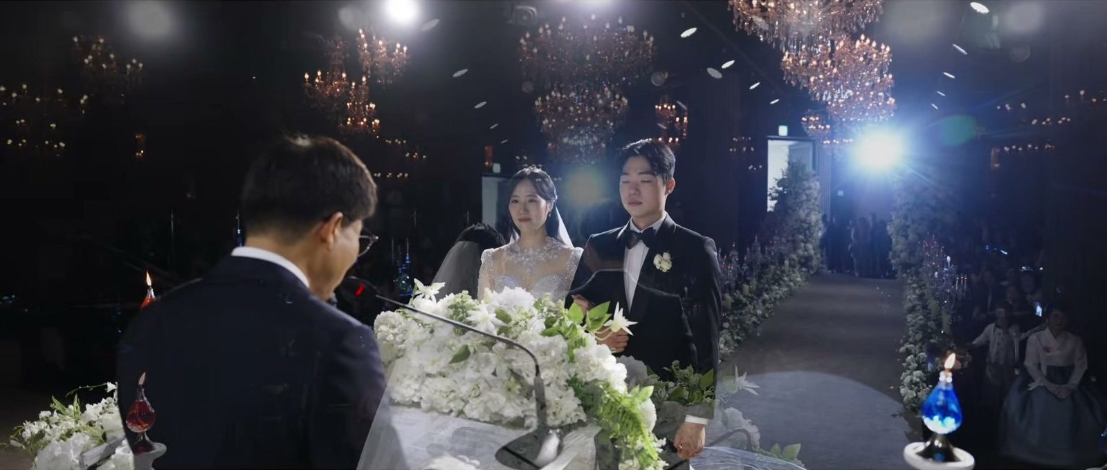 단상 마이크 앞에 선 사람의 뒷모습 너머로 신랑 신부와 꽃장식 버진로드, 크리스탈 샹들리에가 걸린 어두운 천장이 보이는 장면