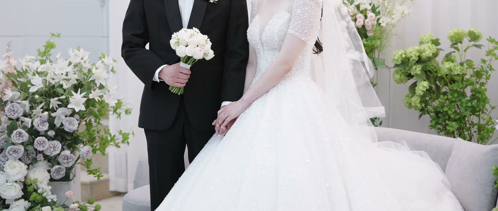 회색 소파와 흰색·연보라색 꽃장식 앞에서 흰 꽃 부케를 든 신랑과 손을 맞잡은 신부