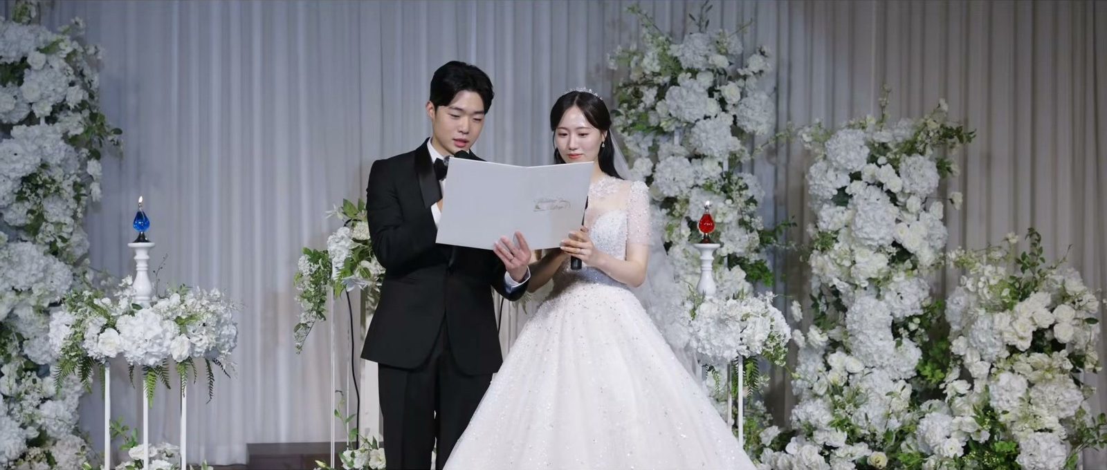 흰 커튼과 흰 꽃장식 앞 단상에서 서약을 읽는 신랑 신부, 양옆 받침대 위의 파란색·빨간색 유리 촛불
