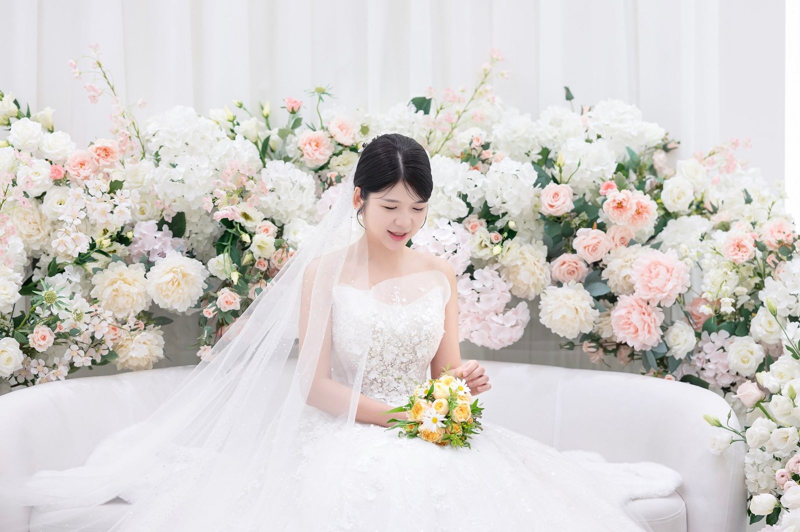 분홍과 흰 꽃 장식 앞 흰 소파에 앉아 노란 꽃 부케를 내려다보는 신부