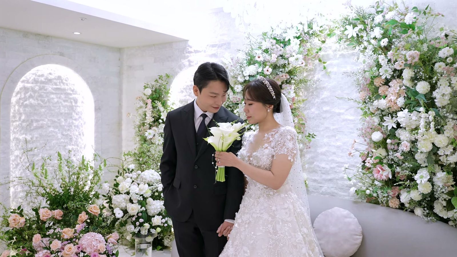흰 돌벽과 꽃 장식 앞에서 신랑 신부가 흰 칼라 부케를 함께 내려다보는 신부대기실