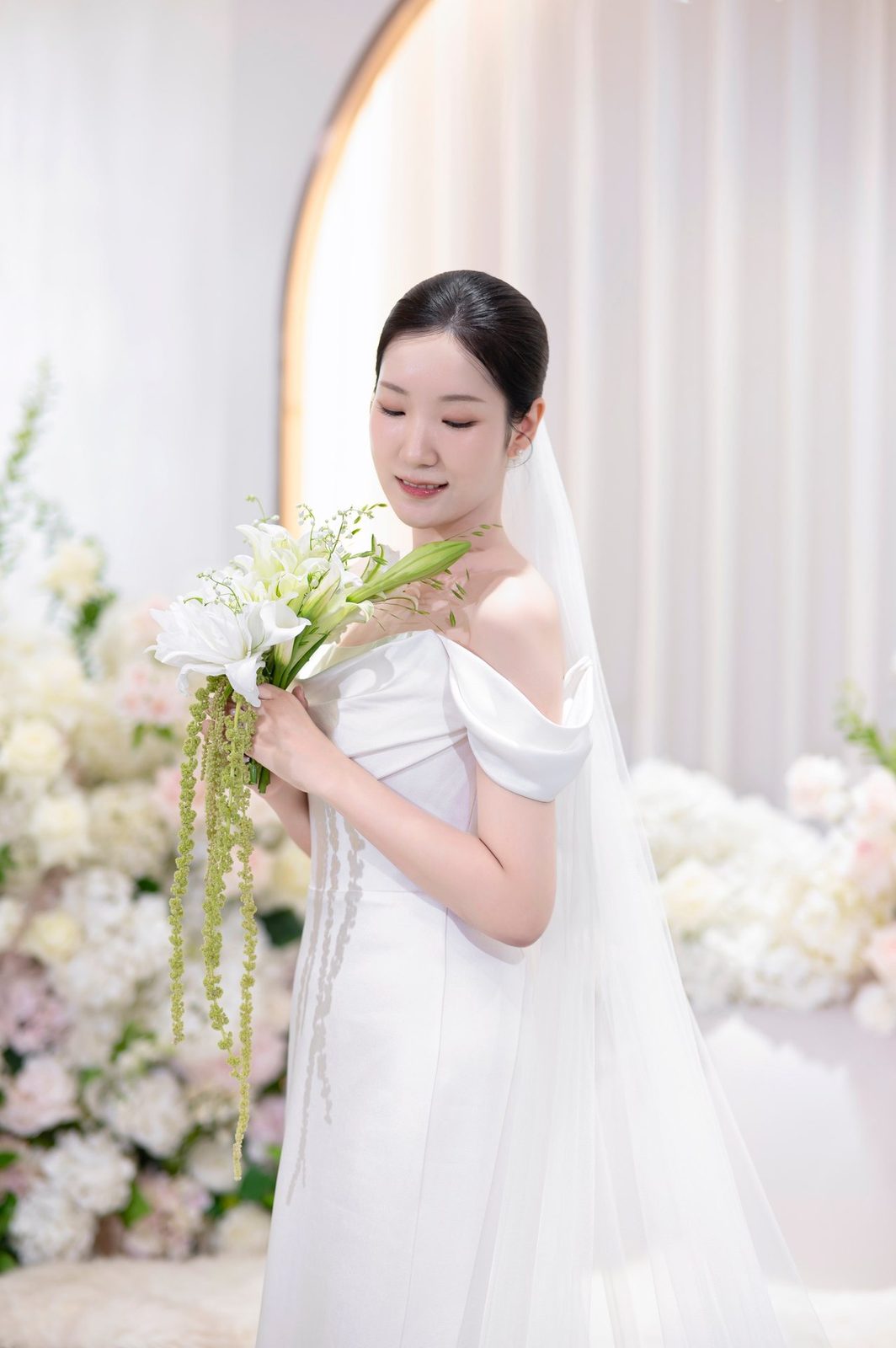 아치 테두리와 흰 커튼 앞, 흰 꽃 더미 사이에서 백합 부케를 내려다보는 신부