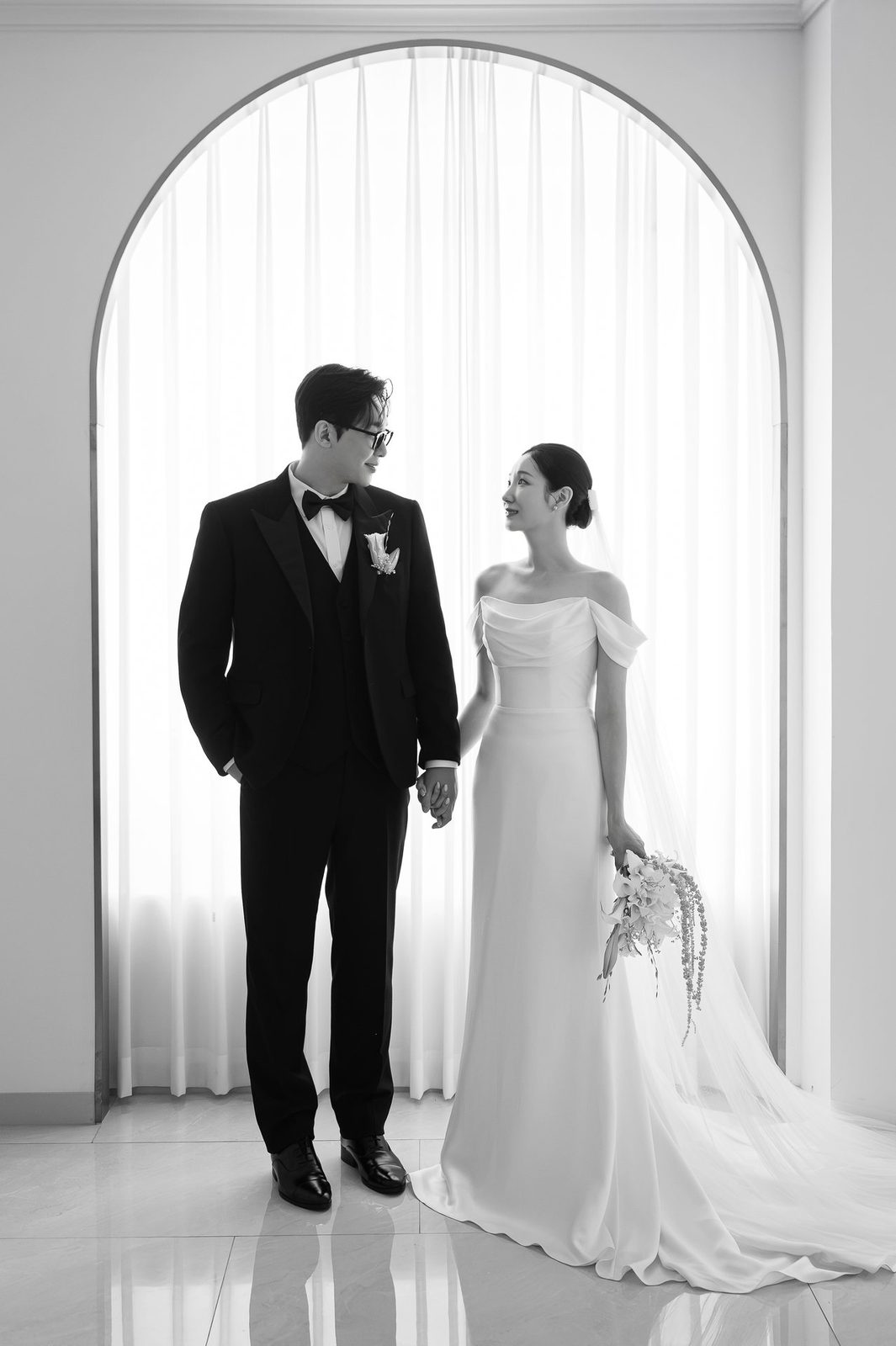 흰 커튼이 드리운 아치형 창 앞에서 손잡고 서로 바라보는 신랑 신부의 흑백 사진