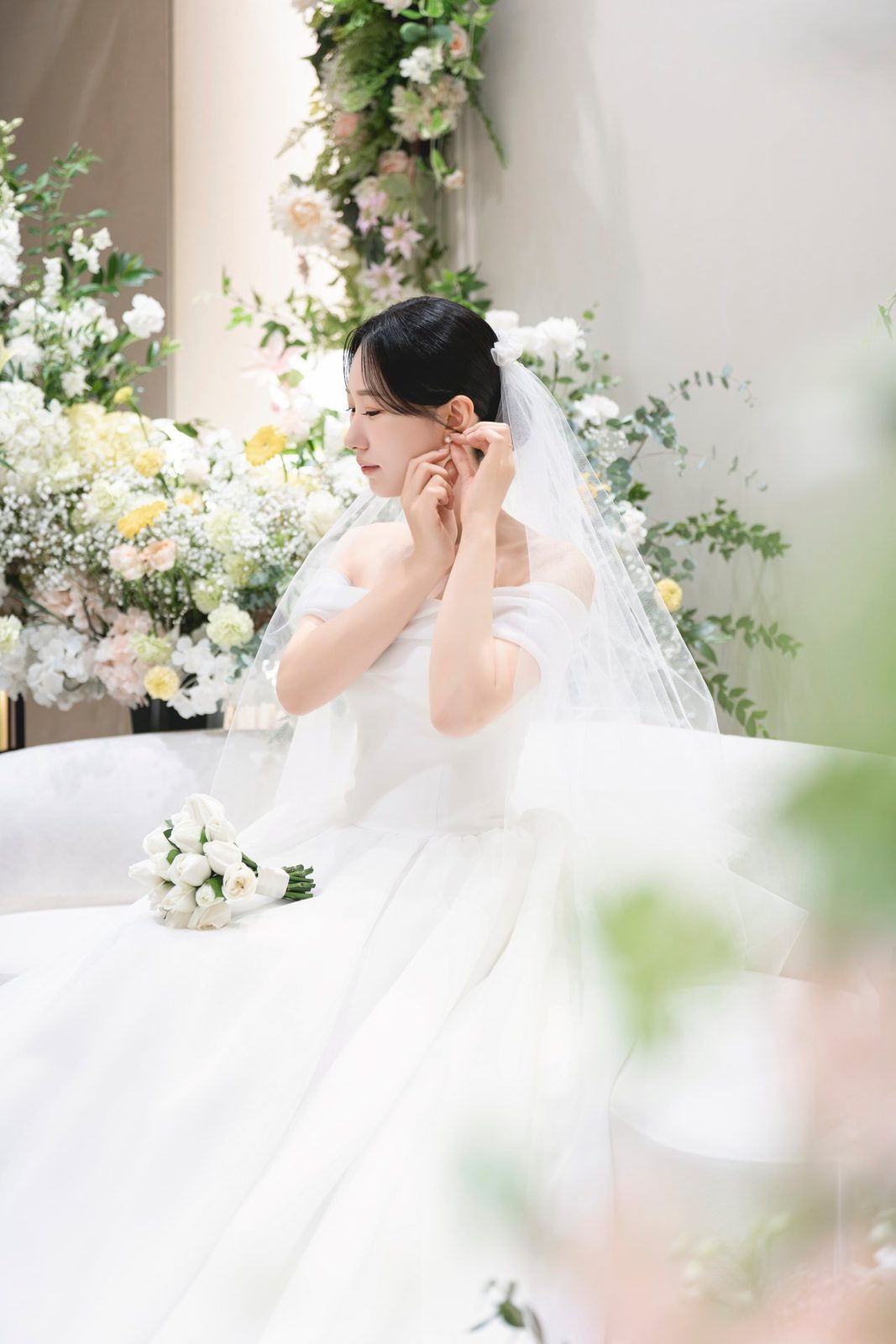 흰 소파에 앉아 귀걸이를 매만지는 신부, 뒤로 흰 꽃과 초록 잎 장식